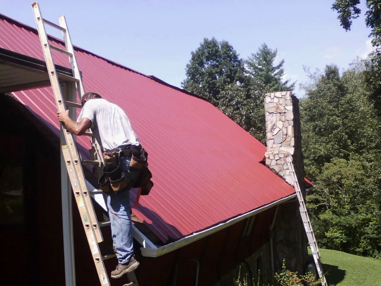 Roofer on a ladder beside a red metal roof and stone chimney — Soffit & Fascia Repair by Roofing Brothers in Cumberland Gap